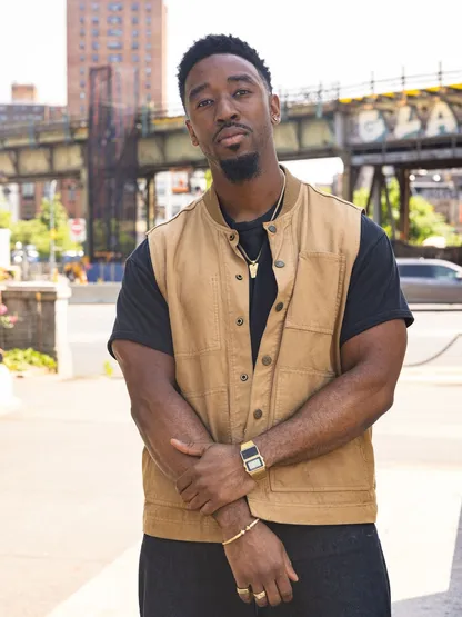 Portrait of Brandon, standing in a work vest on a street under an elevated rail bridge.